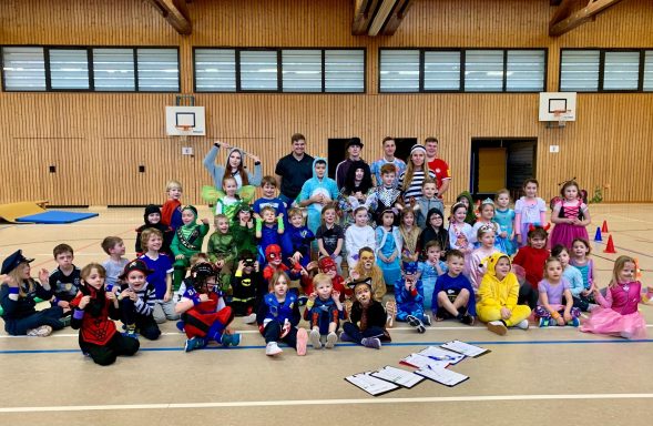 Gruppenselfie von Kindern in bunten Kostümen in einer Sporthalle.
